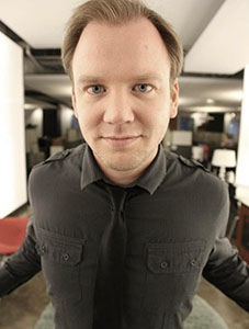 A man with light hair and blue eyes wearing a dark grey shirt and tie, looking directly at the camera.