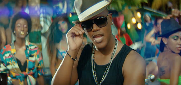 A man wearing a fedora, sunglasses, and beaded necklaces poses in front of a group of people at a party.