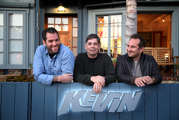 Three men leaning on a blue wooden fence with the word KEVIN printed on it in front of a house.