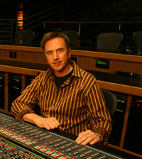 A man in a striped shirt sits behind a large professional audio mixing console in a dark studio.