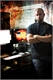 A man with a beard stands with his arms crossed in a dimly lit room with computer monitors.