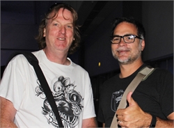 Two men, one with long blond hair and a white t-shirt with a graphic, the other with short dark hair and glasses, give a thum