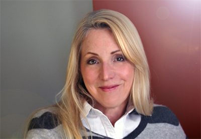 A blonde woman with a slight smile wearing a white collared shirt and a grey striped sweater.