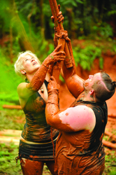 Two people covered in brown mud playing with a long wooden pole in a forest.