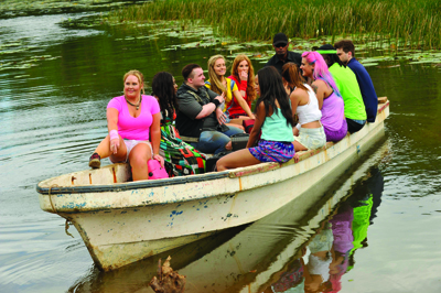 A group of young adults sitting together in a small white rowboat on a calm lake near tall grass.