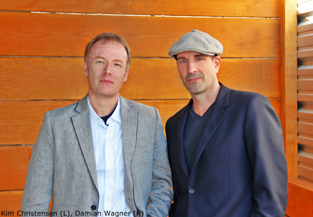 Kim Christensen in a grey blazer and Damian Wagner in a dark suit and grey cap standing against wood paneling.