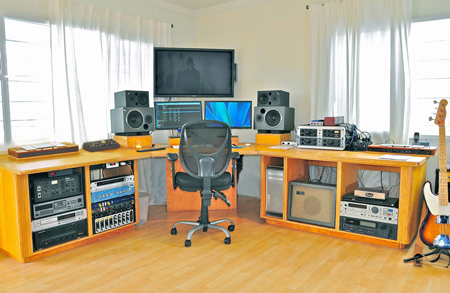 A music production studio featuring a desk with computer monitors, studio speakers, rack-mounted audio gear, and a chair.