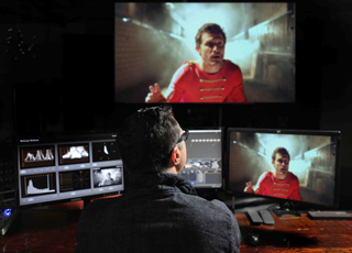A person sits before three computer monitors displaying video editing software and footage of a man in red.
