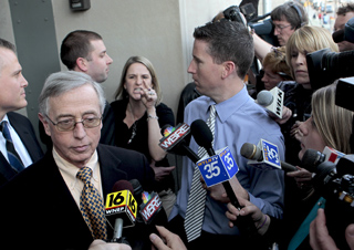 A man in a suit and glasses speaks to a group of reporters holding various news microphones.