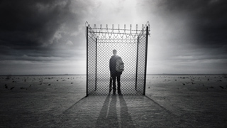 Silhouette of a person standing in front of a tall chain-link fence under a dark, cloudy sky.