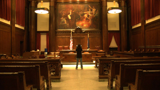 A person stands in the center aisle of a large, wood-paneled courtroom facing a judge's bench.