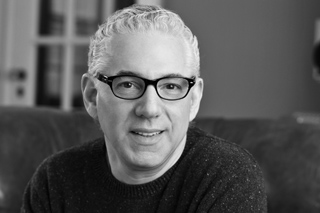 Black and white headshot of a middle-aged man with short wavy hair and dark-rimmed glasses smiling slightly.
