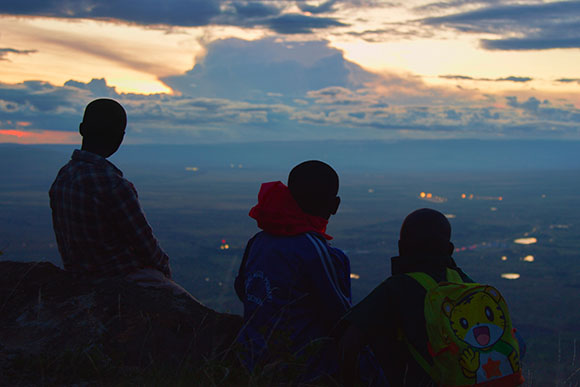 Silhouettes of three people sitting on a hill overlooking a valley at sunset.