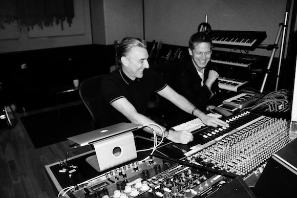 Two men smiling while operating a large audio mixing console in a recording studio.