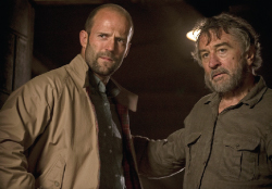 A bald man in a tan jacket looks intensely at an older, bearded man in a dark shirt.