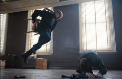 A man in dark clothing jumps mid-air holding a wooden chair toward a man crouched on the floor.