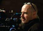 A bald man with sunglasses on his head looks to the side next to a professional film camera.