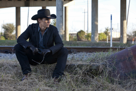 A man wearing a black cowboy hat, dark suit, and leather gloves sits on dry grass near railroad tracks.