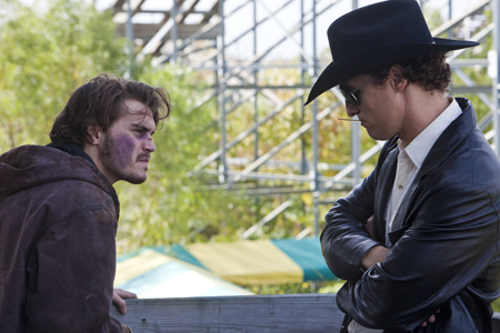 A man with a bruised face stares at a man in a black cowboy hat and leather jacket.