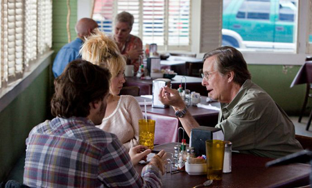 A man in a green shirt talks to a woman and man at a diner table.