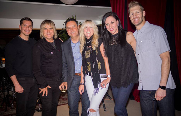 Six smiling people standing in a row, posing for a group photo in a dimly lit room.