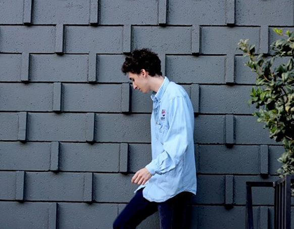 A young man in a light blue button-down shirt walks past a dark grey geometric textured wall.