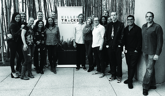 A group of twelve people posing together outdoors in front of a Killer Tracks banner and bamboo.