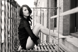 Black and white photo of a woman in a dark polka dot shirt sitting on a wooden balcony ledge.