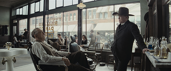 A man in a black suit stands near a counter in a vintage barbershop with large windows and patrons.