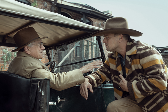 A man in a tan jacket sits in a vintage car while a man in a patterned coat leans in to speak with him.