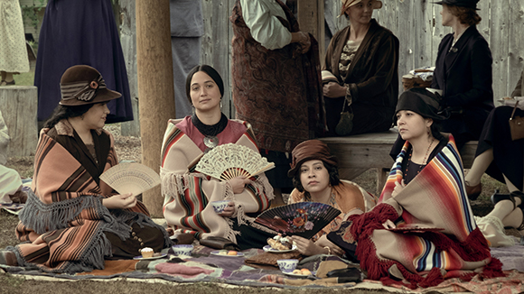 Several women in early 20th-century attire sit on a blanket with fans and tea.