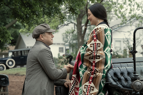 A man in a grey suit kneels to hold the hands of a woman wrapped in a colorful geometric patterned blanket.