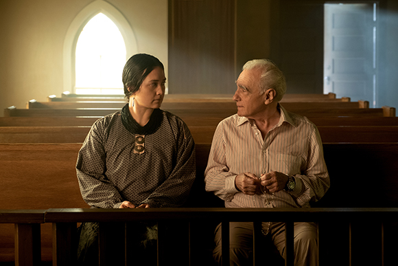 A young woman in dark clothing and an older man in a striped shirt sit in wooden pews facing each other.