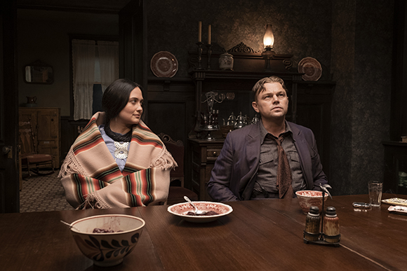 A woman wrapped in a striped blanket and a man in a suit sit at a dark wooden table in a vintage room.