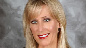 A blonde woman with blue eyes and red lipstick smiling against a gray background.