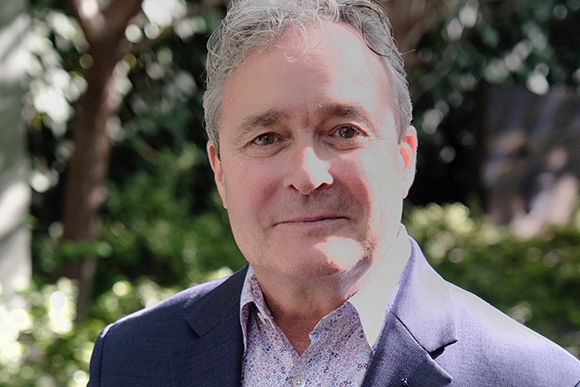 A middle-aged man with graying hair wearing a blue blazer and patterned shirt, smiling slightly outdoors.