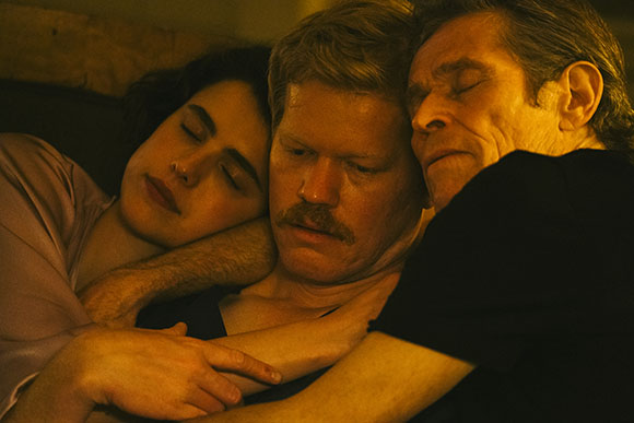 Three people with eyes closed, embracing closely in warm, dim lighting.