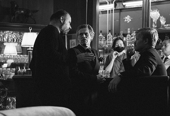 Four men in dark suits converse in a dimly lit bar with glassware in the background.
