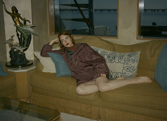 A woman in a mauve tunic reclines on a tan sofa with patterned pillows near a window.