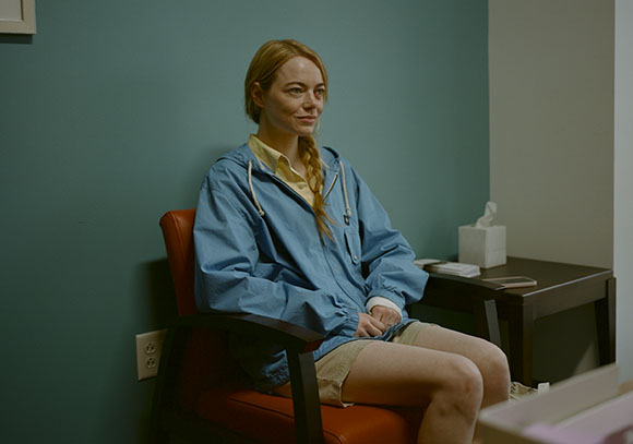 A woman with blonde hair sits in a red chair wearing a blue jacket and tan shorts.