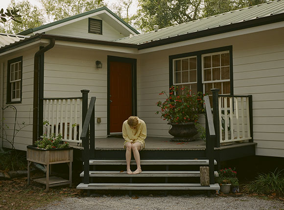 A person in a yellow robe sits on the edge of wooden porch steps in front of a white house.