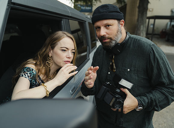A woman leans out of a car window toward a man holding a professional camera and wearing headphones.