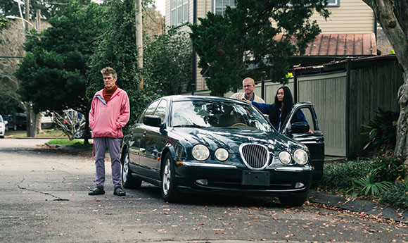 A man in a pink jacket stands by a dark sedan while two others stand near the open passenger door.