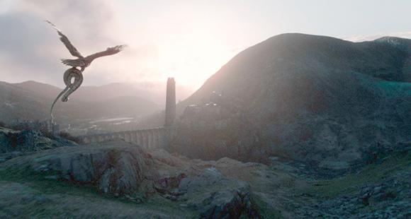 A dragon flies through a misty sky over rugged mountains and a tall stone tower at sunset.