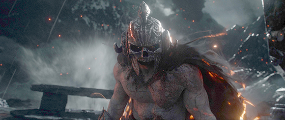 A muscular warrior wearing a silver skull-shaped helmet and dark cape in a snowy, smoky environment.