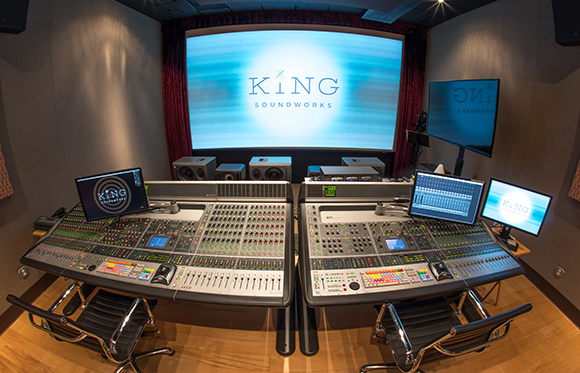 Two large audio mixing consoles sit in a studio facing a large projection screen displaying a logo.