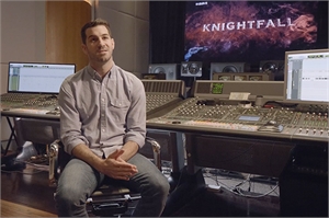 A man in a light blue shirt sits in a chair in front of a large mixing console in a studio.