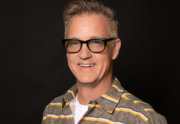 A smiling man with short wavy hair and black-rimmed glasses wearing a brown, yellow, and white striped shirt.