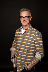 A smiling man with glasses and tattoos wearing a yellow and grey striped button-down shirt.