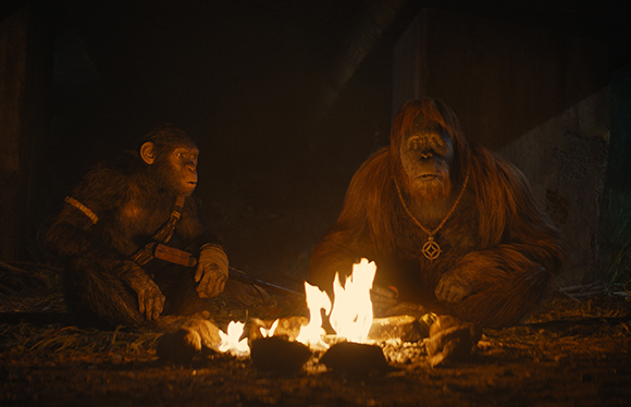 Two primates sit on the ground around a small, glowing campfire in a dark, dimly lit environment.